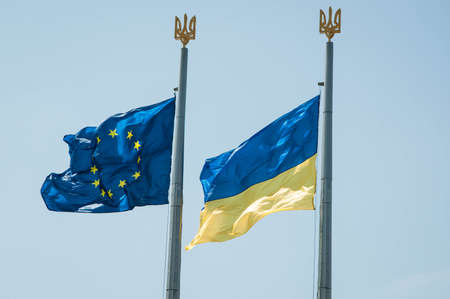 Ukrainian and European union flag waving on blue sky with coat of arms of Ukraine.
