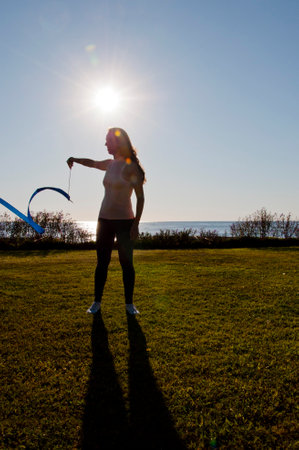 Gymnastics flexibility. Flexible girl in summer sea. Woman silhouette with gymnastics ribbon. Rhythmic gymnast performing yoga. Sport warm up motivation. Gymnast hold ribbon in backlit. Ribbon dancing.の写真素材
