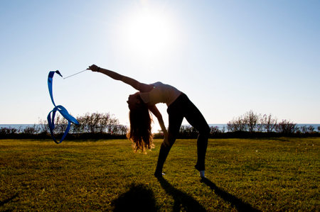 Flexible girl in summer sea. Woman silhouette with gymnastics ribbon. Sport flexibility. Gymnast hold ribbon in backlit. Gymnastics flexibility.の写真素材