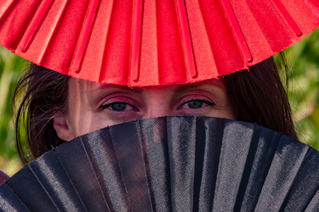 Cultural folk elegance hidden face. Geisha girl. Secret gaze of dancer. Eyes of mystery feminine strength. Mysterious eyes behind traditional fans. Flamenco dancer. Woman eyes red black fan. See look.の写真素材