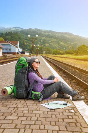 Happy girl tourist wearing backpack, sits on a platform waiting for a train. Traveler is waiting at the railway station. Travel concept. Tourist on railroadの写真素材