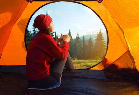 View from inside a tent on mountains landscape. Camping concept. Sunset inside a tent. Young woman sitting in the tent with cup looking at the mountain landscapeの写真素材