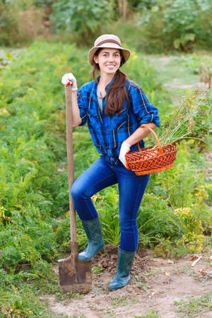 autumn harvesting concept. Happy smiling caucasian female farmer or gardener holding spade and basket of harvested vegetables . Woman working in the gardenの写真素材