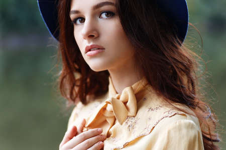 Outdoor closeup fashion portrait of glamour sensual young stylish lady wearing trendy hat. Autumn woman. Girl portrait. Fashion woman with curly hair and floppy hat. Closeup portrait of young beautiful brunette girl with brown eyes. Looking at cameraの写真素材