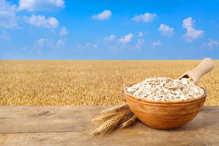 Oat flakes in bowl. Ears of oats and oatmeal in bowl on table with field on the background. Golden field and blue sky. Uncooked porridgeの写真素材
