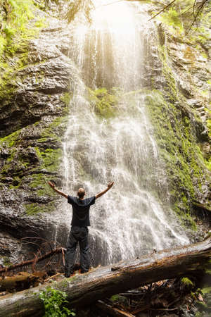 man embracing waterfall with open arms. Tourist looking at the waterfall. Freedom, happiness, hiking concept. Back view of man standing in front of waterfall with outstretched handsの写真素材