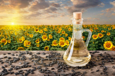 sunflower oil in glass bottle and scattered seeds on wooden table with blooming field on the background. Sunflower field on sunset. Photo with copy space. Agriculture and harvest conceptの写真素材