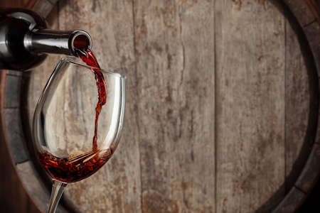 red wine pouring from bottle into glass with old wooden barrel as backgroundの写真素材