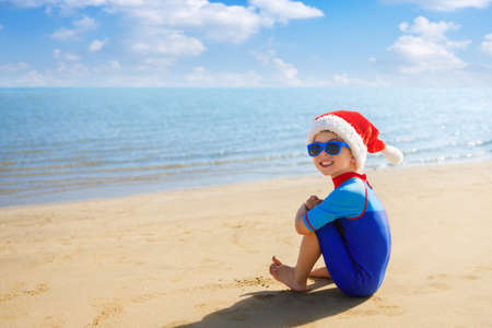 smiling child boy in santa hat, sunglasses and swimsuit sitting on the sand beachの写真素材