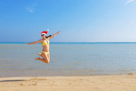 happy woman in santa hat and sunglasses jumping on sand beachの写真素材