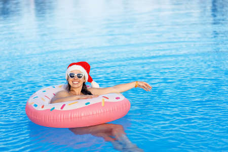 attractive young woman in santa hat and sunglasses relaxing on inflatable ring in swimming poolの写真素材