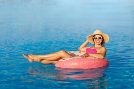 smiling young woman in straw hat and sunglasses relaxing on inflatable ring in swimming pool
