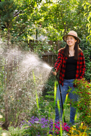 smiling young woman gardener watering flowers and plants in garden with hoseの写真素材