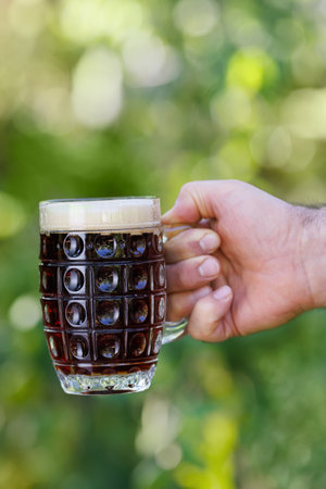 closeup male hand holding mug of dark beer with foam and making toast on green natural backgroundの写真素材