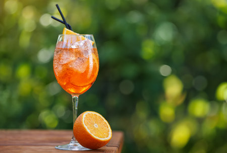 glass of cold aperol spritz cocktail with straws and orange on wooden table with green natural backgroundの写真素材