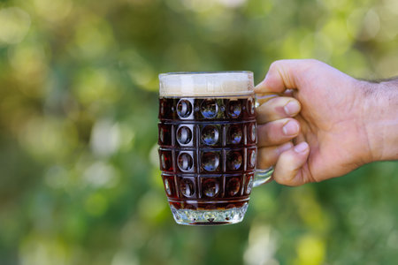 closeup male hand holding mug of dark beer with foam and making toast on green natural background