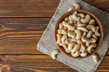 Wooden bowl holding raw cashew nuts sitting atop textured linen cloth against dark wooden surface,...