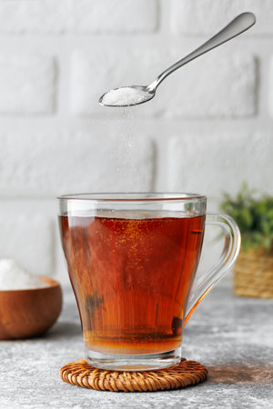 White sugar falling from teaspoon into glass cup of hot tea placed on wicker coasterの写真素材