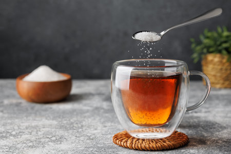 White sugar falling from teaspoon into glass cup of hot tea placed on coaster against grey backgroundの写真素材