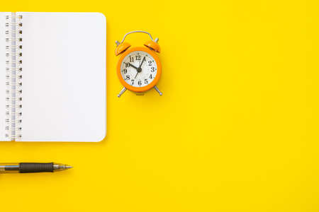 Top view of a notebook, a black pen and a small orange retro-style alarm clock. Isolated on yellow background with lot of copy space on the right.の写真素材