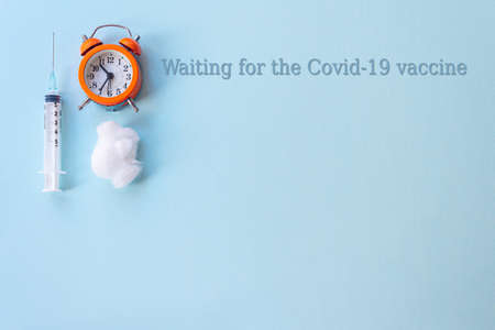 Concept image with text Waiting for the  vaccine. Syringe, cotton ball and orange alarm clock on a...