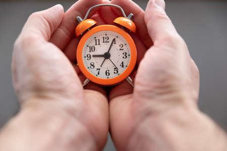 Caucasian white man hands holding a small orange alarm clock in the palm. Concept of time.の写真素材
