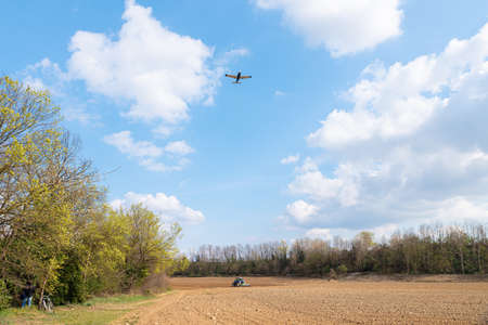 Collegno, Italy. An airplane flies over the countryside while a tractor is plowing the land of a field.の写真素材