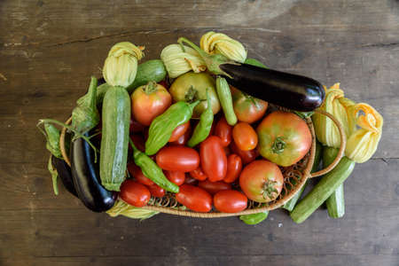 Overhead shot of a woven basket on a wooden table filled with ripe organic tomatoes and other vegetables.の写真素材