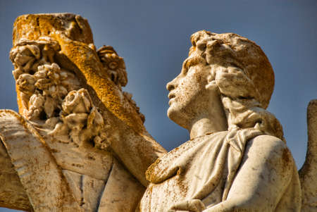 A stone statue of an angel touches a garland on a cross.