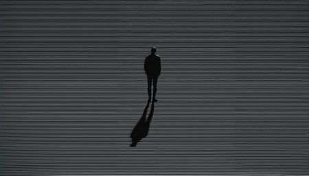 A stark black and white image showing a solitary human figure in silhouette, standing against a background of perfectly horizontal parallel lines. The figure casts a long, dramatic shadow that stretches diagonally across the composition, creating a striking contrast with the geometric precision of the lined background. The minimalist style and strong geometric elements give the image a sense of isolation, contemplation, and artistic sophistication.の素材