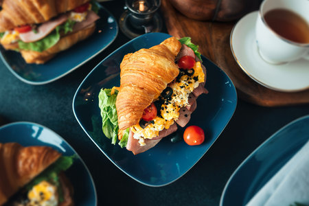 Fresh croissant sandwich with ham, scramble egg, tomato with tea on table, top viewの写真素材