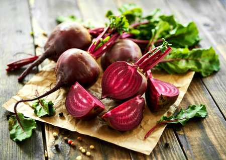Whole and sliced beetroots with vibrant red flesh are arranged on brown paper atop a dark wooden surface, accompanied by fresh green leaves and peppercorns.の写真素材