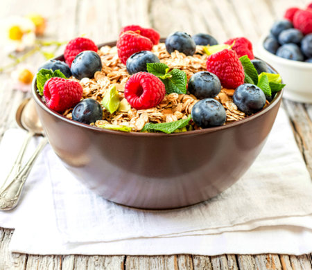 A brown bowl filled with oatmeal topped with fresh raspberries, blueberries, and mint leaves. The bowl sits on a white napkin on a wooden surface.の写真素材