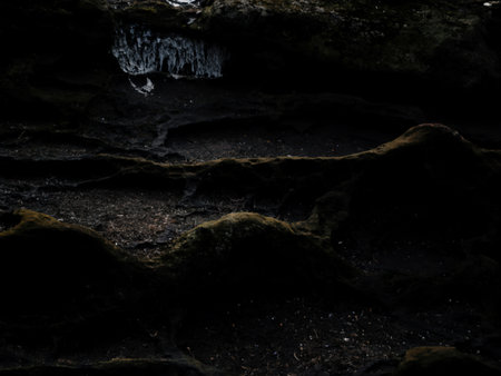 A close-up image reveals dark, moss-covered rocks with a small waterfall cascading in the background. The texture is rough and uneven, showcasing the natural erosion of the stone.の写真素材