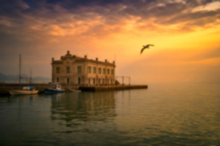 A serene sunrise illuminates a historic building situated on a calm harbor, with small boats gently...