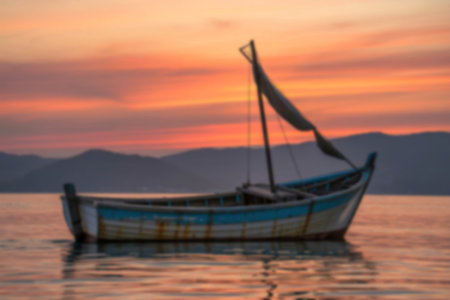 A rustic, weathered sailboat gently floats on calm water, silhouetted against a breathtaking sunset over distant mountains. The scene evokes tranquility and peace.の写真素材