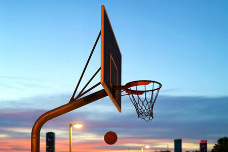 A basketball hangs in the air, about to enter the hoop of an outdoor court at sunset. The urban...