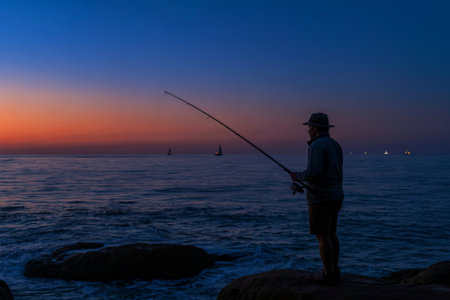 A lone fisherman stands on a rocky outcrop, silhouetted against a vibrant sunset, casting his line into the ocean. Sailboats are visible on the horizon.の写真素材