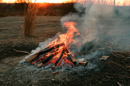 A vibrant bonfire crackles and smokes in a field at sunset. The flames are bright orange and yellow, casting a warm glow against the darkening landscape.の写真素材