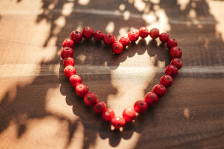 A heart shape is formed by small, red berries on a brown wooden surface. Sunlight casts shadows on the wood.の写真素材