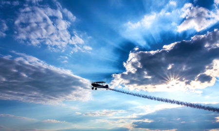 A biplane leaves a smoky trail as it soars through a partly cloudy sky, sunbeams radiating from...