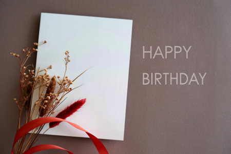 A minimalist birthday card mockup featuring a blank white card adorned with dried flowers, a red ribbon, and the words Happy Birthday subtly displayed on a mauve background.の写真素材