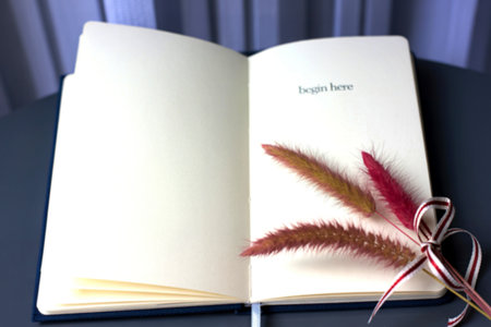 A navy blue notebook lies open, displaying the words begin here. Two sprigs of dried flowers, tied with a ribbon, rest on the open pages.の素材