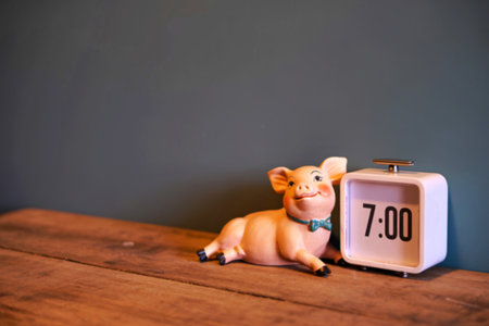 A charming pink pig figurine with a bow tie rests beside a white retro flip clock displaying 7:00 on a rustic wooden surface against a muted green background.の素材
