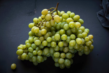 A close-up, overhead view of a large bunch of ripe green grapes, artfully arranged on a dark,...