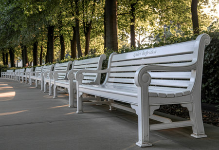 A line of pristine white park benches with ornate details stretches along a paved walkway. Sunlight filters through the dense green trees overhead, casting dappled shadows.の写真素材