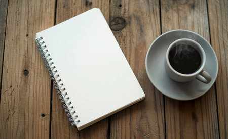 A steaming cup of dark coffee rests on a saucer beside an open, blank spiral-bound notebook. The scene is set on a textured, weathered wooden surface, evoking a sense of calm and productivity.の素材