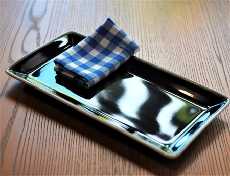 A glossy black rectangular serving platter is set on a dark wooden table. A neatly folded blue and white gingham napkin rests on the platter.の素材