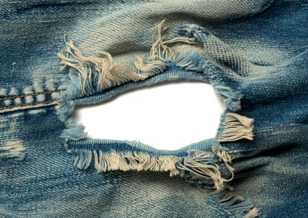 A detailed view of worn blue denim fabric featuring a significant tear with loose, frayed threads and a white background showing through the hole.の写真素材