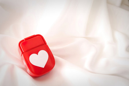 A bright red, heart-shaped pill box rests on smooth, white silk fabric. The luxurious material is softly draped, creating a gentle backdrop for the vibrant red accessory.の写真素材